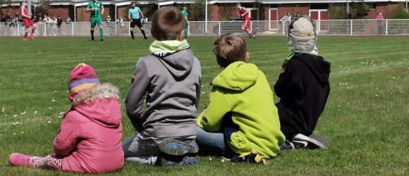 Das Bild zeigt drei Kinder, die am Fußballplatz sitzen